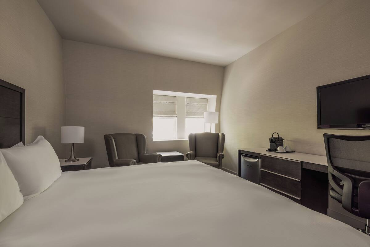 Ein Hotelzimmer mit einem großen Bett, zwei Sesseln, einem Schreibtisch mit Lampe, einem Fernseher und einem Fenster.