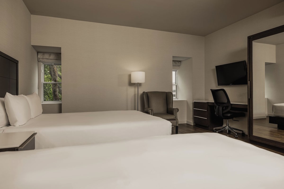 A hotel room with two beds, a desk, a chair, a TV, and a window with a view of trees.