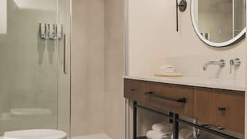 A modern bathroom with a glass-enclosed shower, a white toilet, a vanity with a mirror, and a shelf with folded towels.