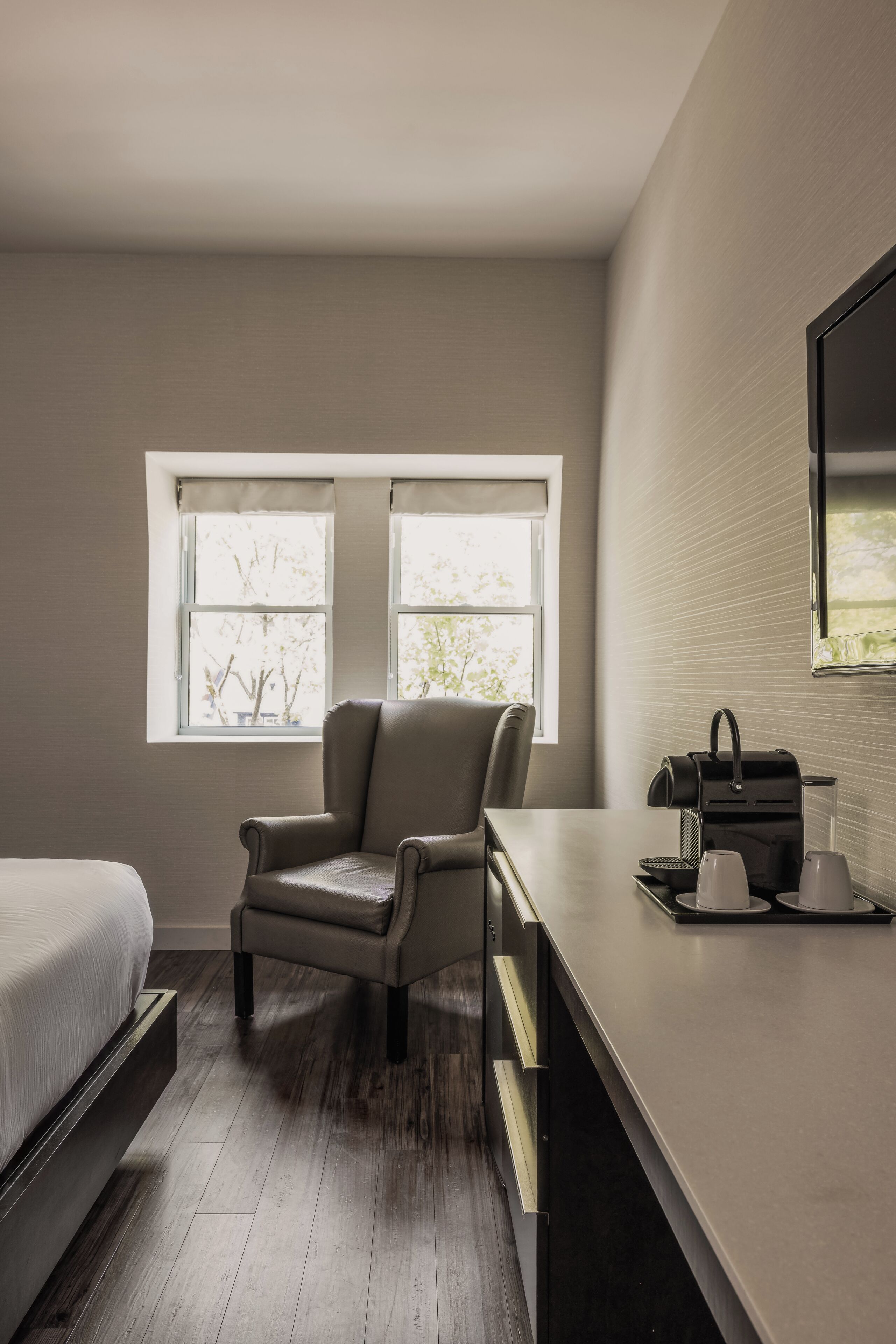 A bedroom with a bed, a chair, a desk, a coffee maker, and a window with a view of trees.