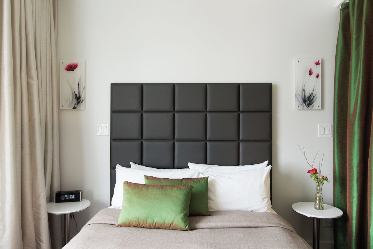 A hotel room with a dark headboard, white pillows, a green pillow, a bedside table with a clock, and artwork on the walls.