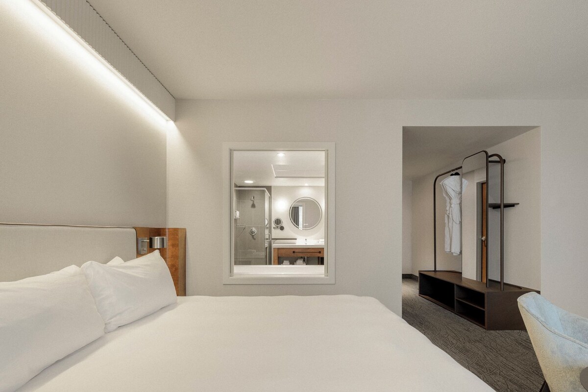 Habitación de hotel con una cama grande, una mesita de noche, un espejo y vistas al baño.