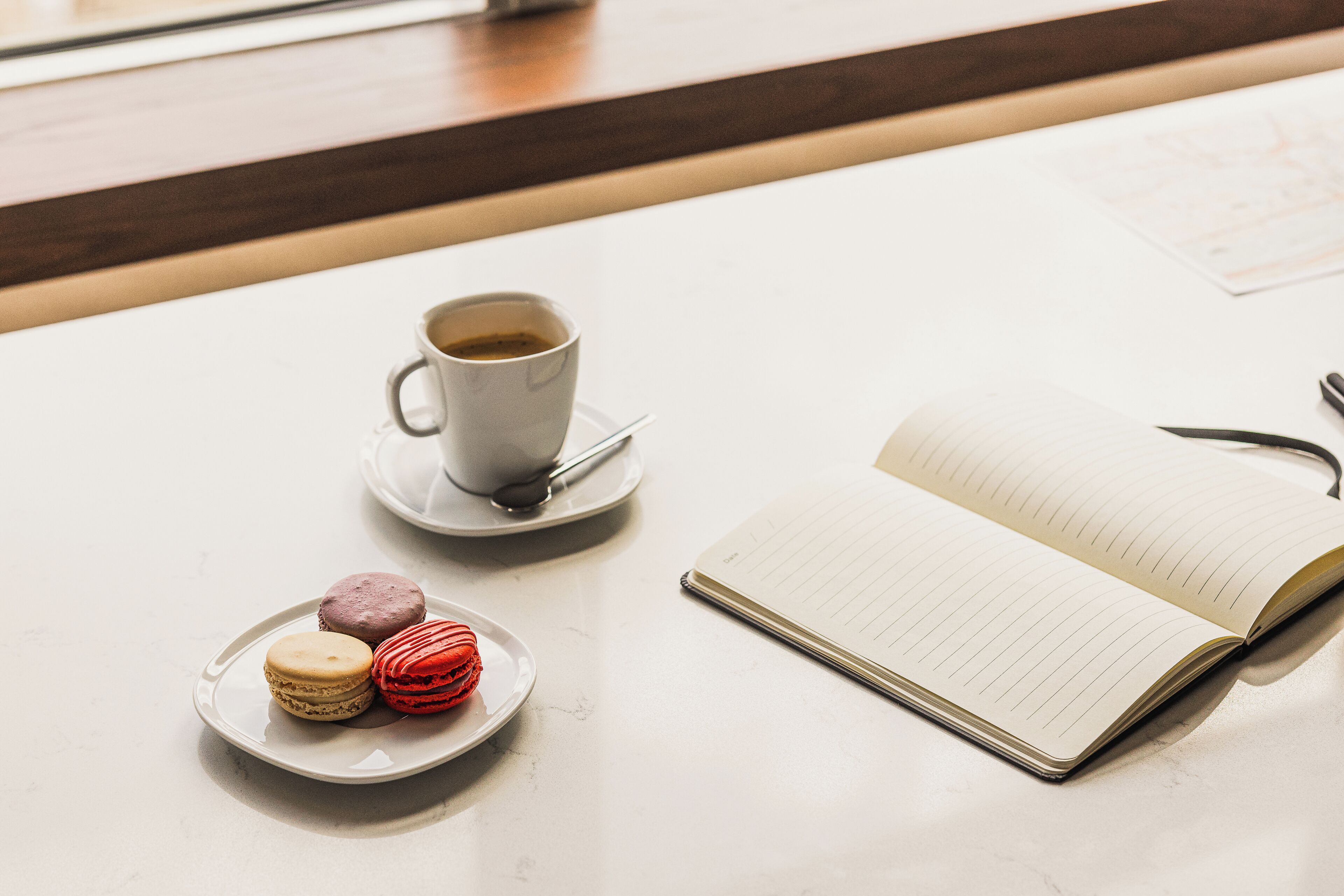 Uma mesa com uma xícara de café, duas travessas de macarons e um caderno aberto.