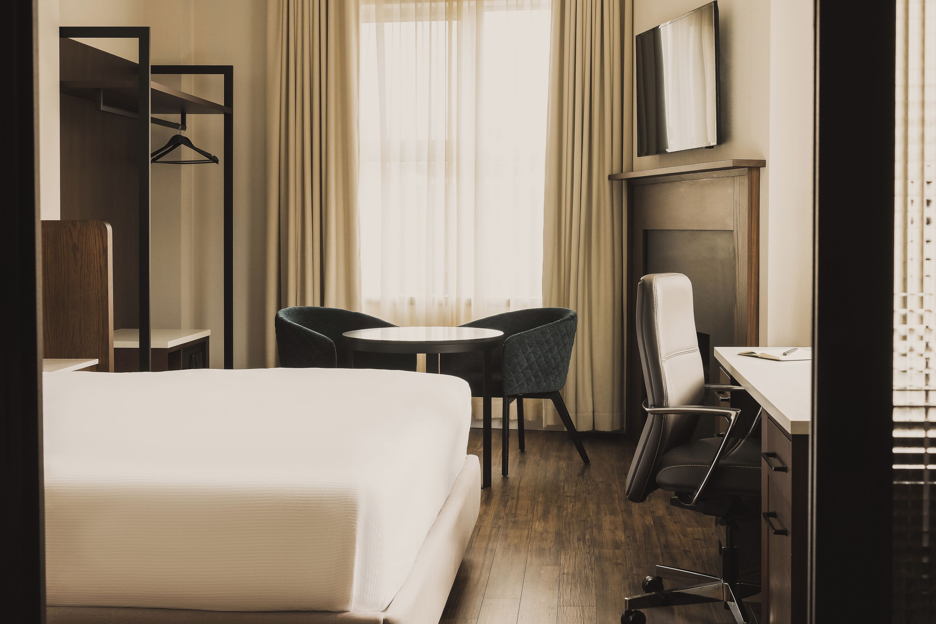 A hotel room with a bed, a desk, two chairs, a TV, and a window with curtains.