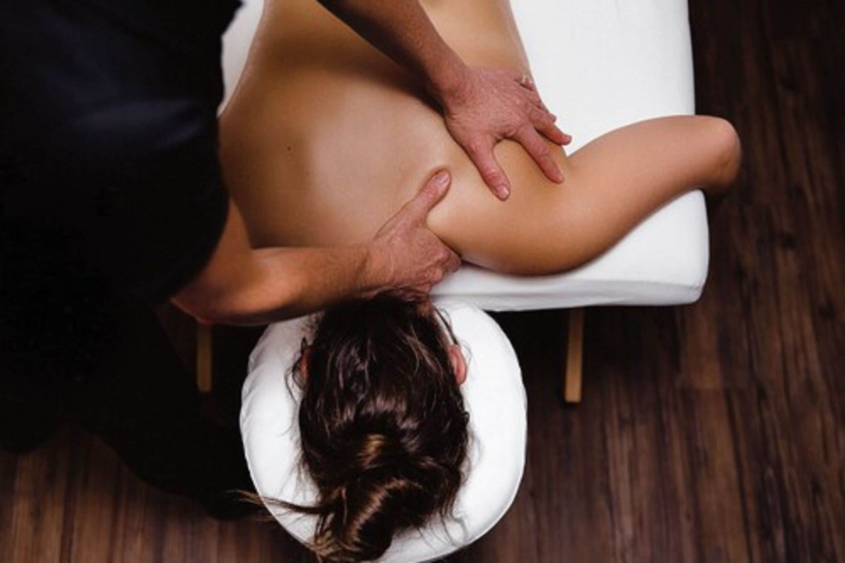 A person receiving a back massage on a white massage table in a room with wooden flooring.