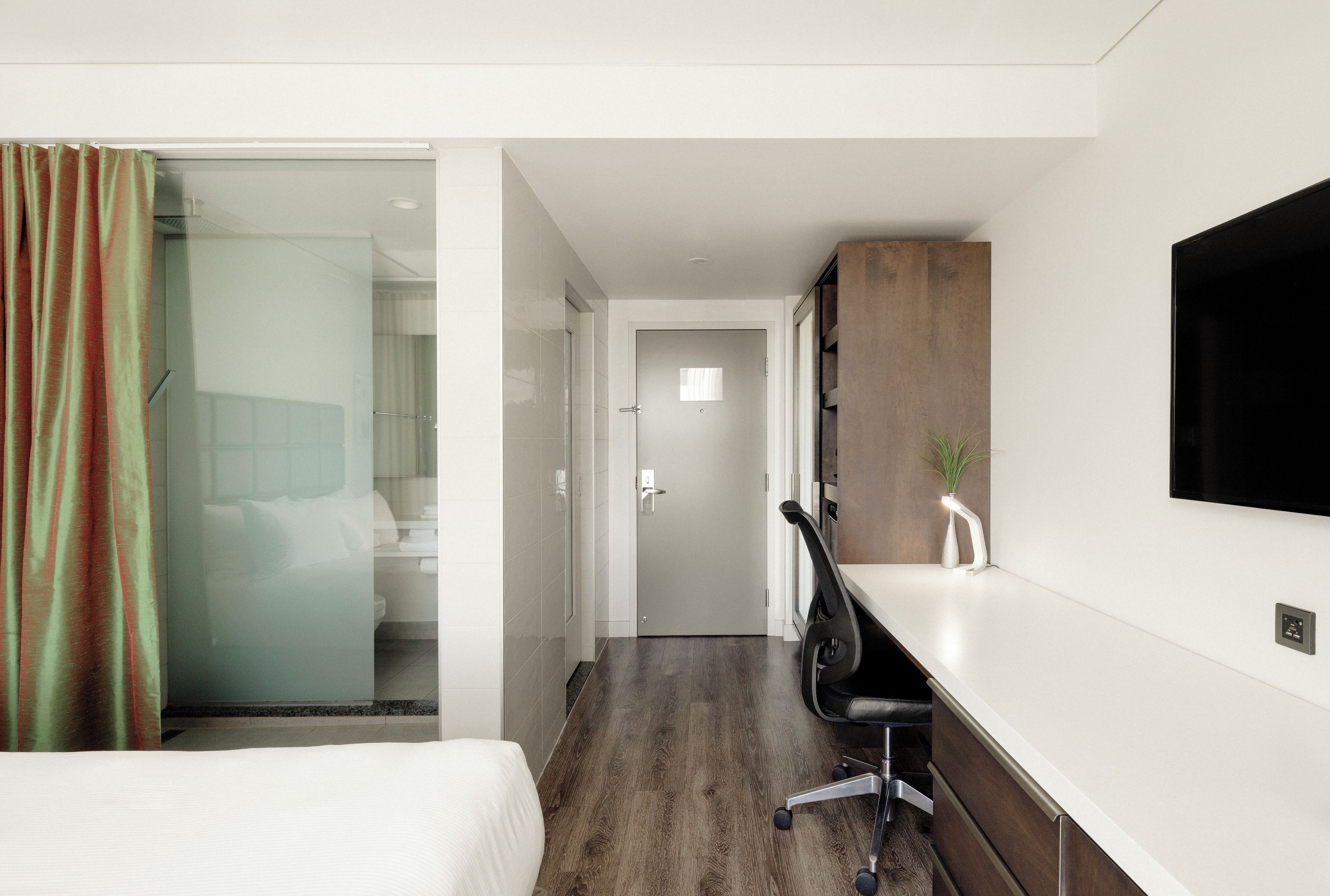 Habitación de hotel con cama, escritorio, silla, televisor y baño visible a través de una puerta de cristal.