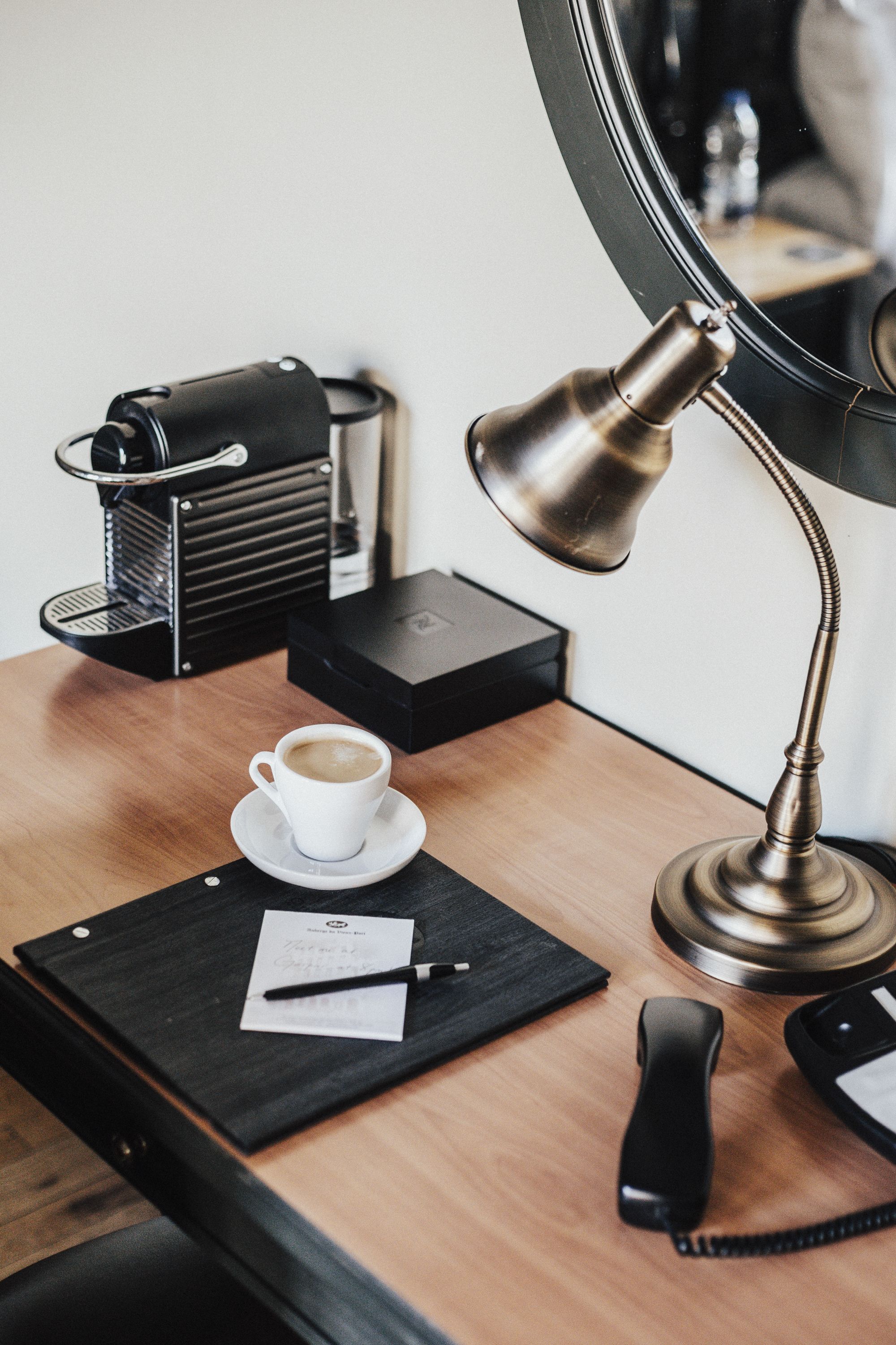 A wooden desk with a coffee cup, a lamp, a telephone, a black notebook, and a pen.