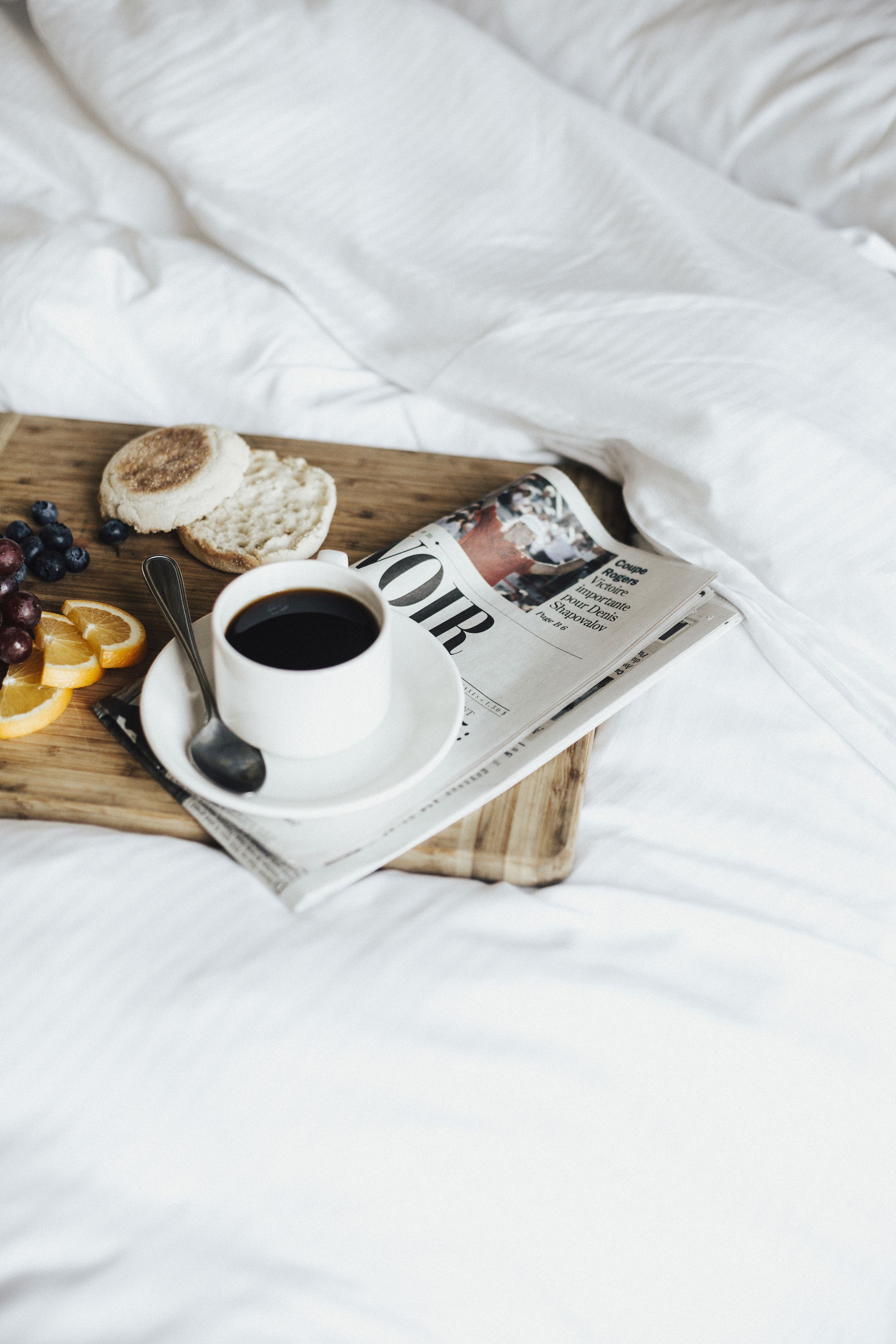A breakfast setup on a bed with a newspaper, a cup of coffee, toast, and fruit.