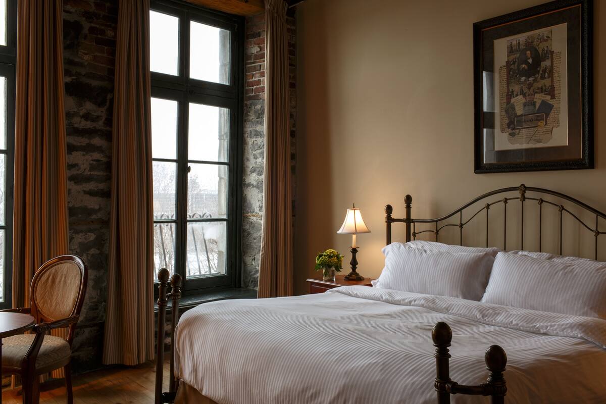 A neatly made bed with white linens, a wooden headboard, and a nightstand with a lamp and a small plant.