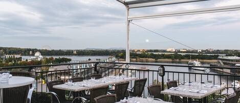 An outdoor dining area with tables and chairs overlooking a river and cityscape.
