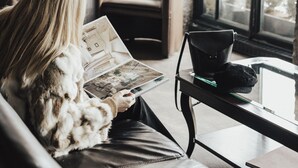 A person reading a book in a room with a leather couch, a chair, a table with a laptop, and a large window with a view of trees. - Auberge du Vieux-Port (Montreal)