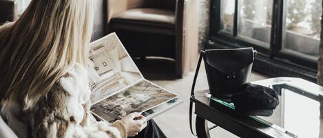 Una persona leyendo un libro en una habitación con un sofá de cuero, una silla, una mesa con una computadora portátil y un amplio ventanal con vistas a árboles.
