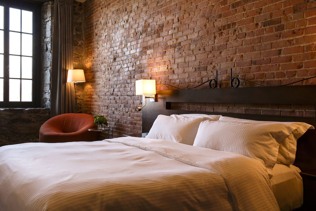 A hotel room with a brick wall, a large bed, a chair, and two bedside lamps.