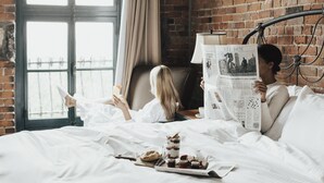 A room with a brick wall, a window with curtains, a bed with white bedding, and a tray with pastries. - Auberge du Vieux-Port (Montreal)