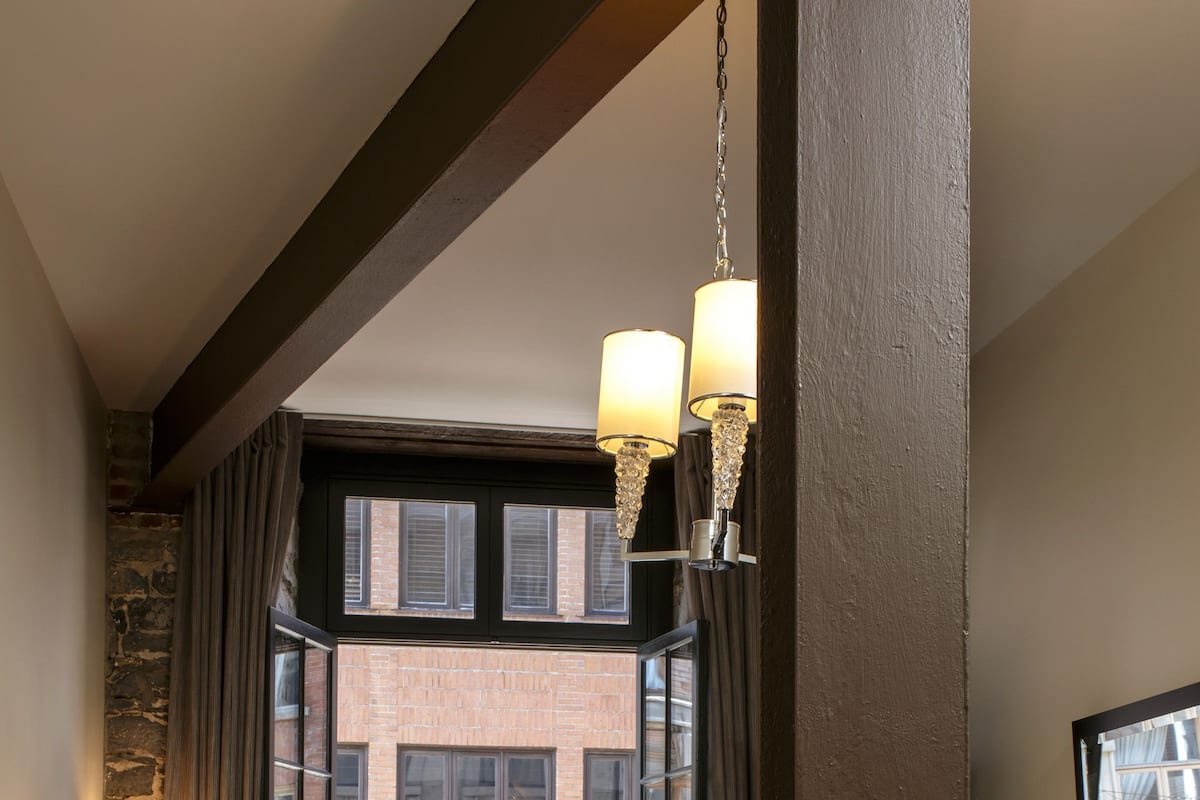 A hotel room with a bed, a nightstand, a lamp, a window with a view of a brick building, and a chandelier.