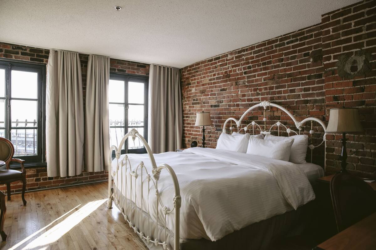 A bedroom with a brick wall, a white metal bed frame, and a view through a window with curtains.
