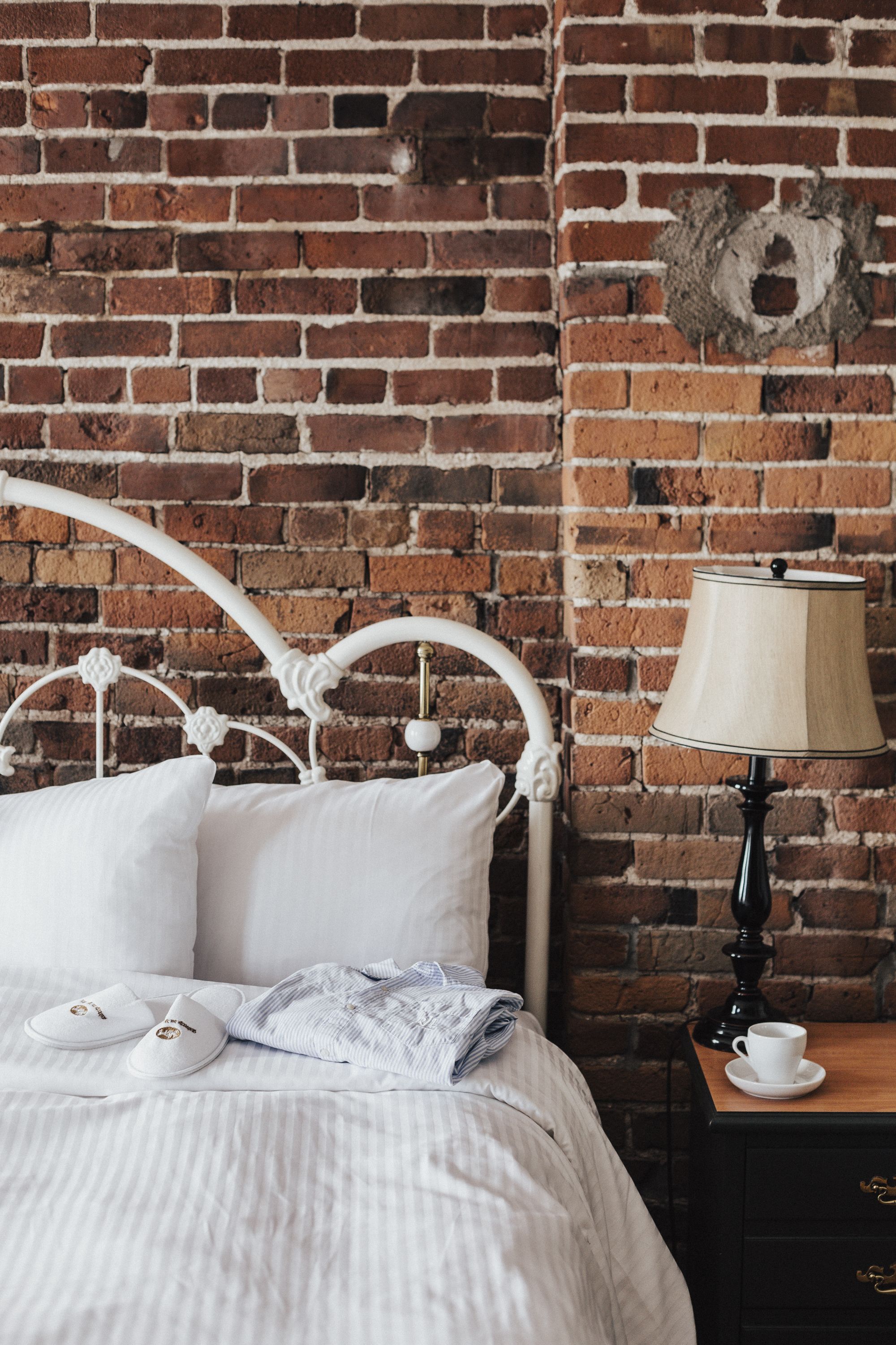 Una cama bien tendida con ropa de cama blanca y almohadas, cabecera de metal blanco, mesita de noche de madera con una lámpara y pared de ladrillo con un plato decorativo.