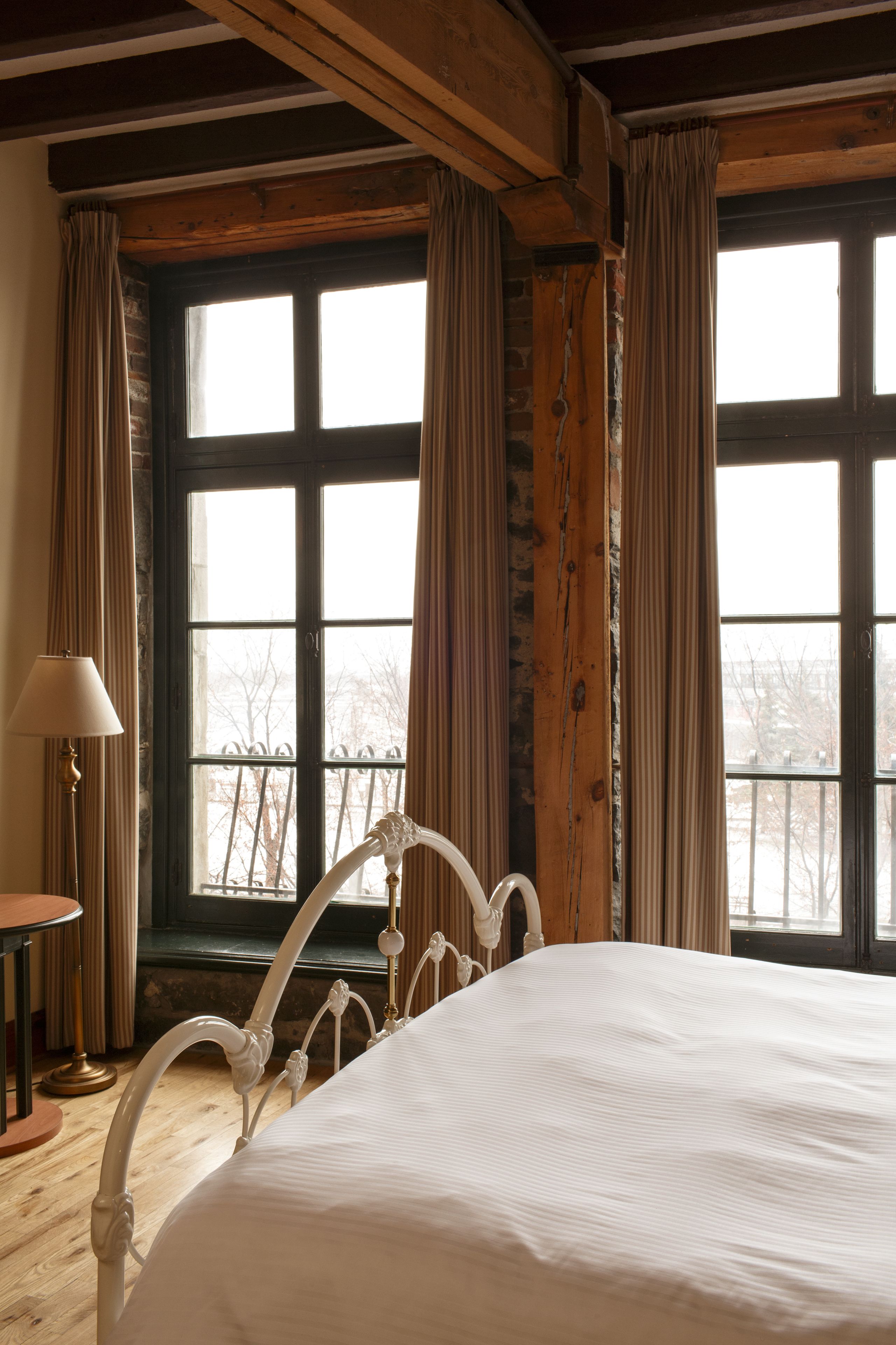 Quarto com uma cama grande, vigas de madeira e vista pela janela com cortinas.