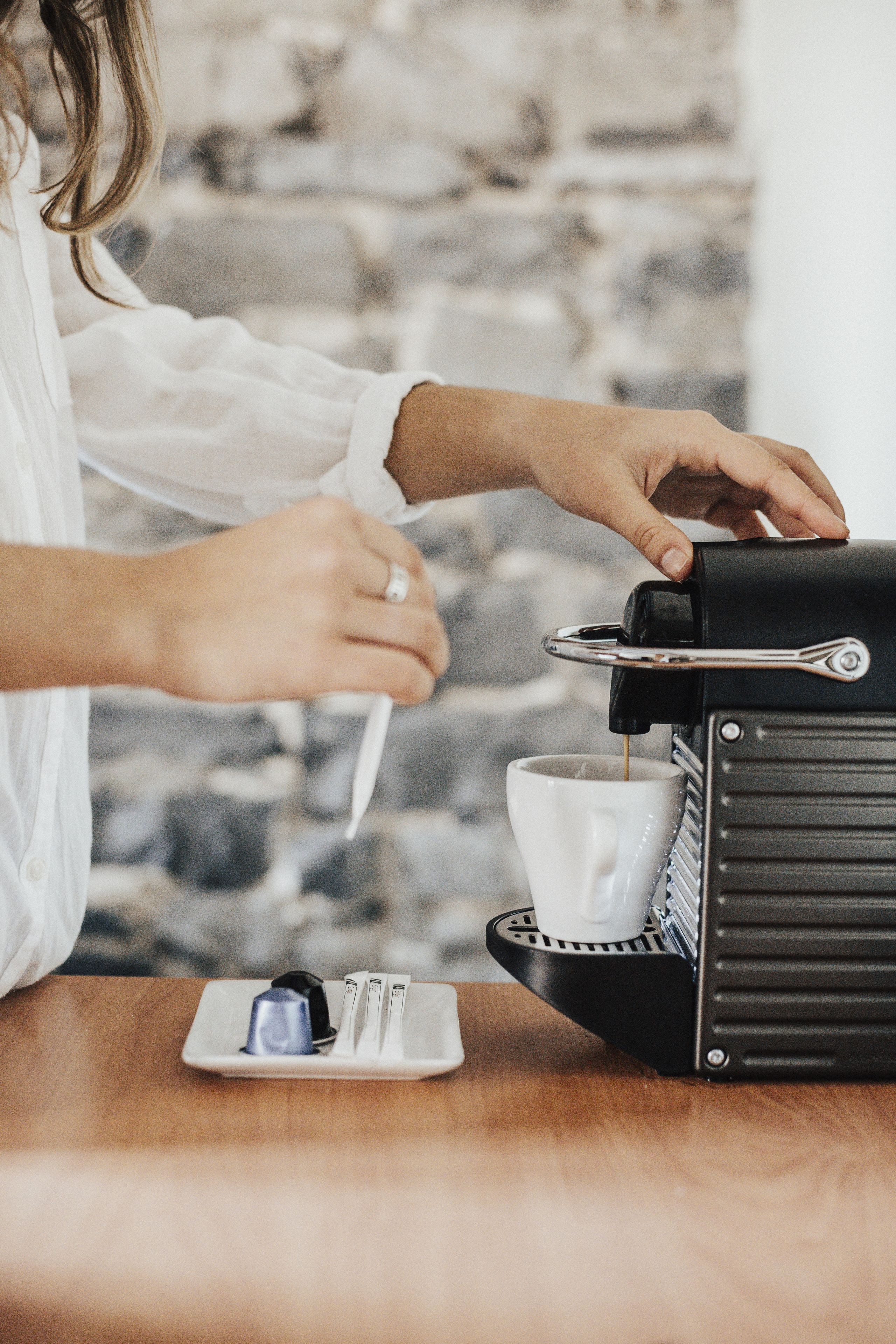 A person operating a coffee machine, with a cup and a plate with a coffee pod and spoon on a wooden table.