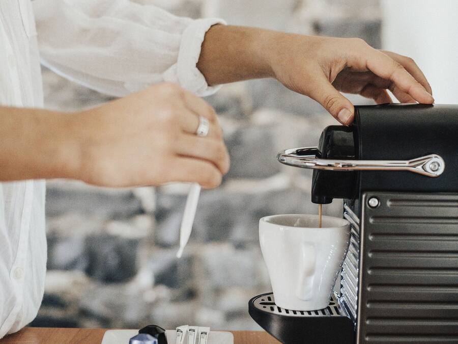 A person operating a coffee machine, with a cup and a plate with a coffee pod and spoon on a wooden table.
