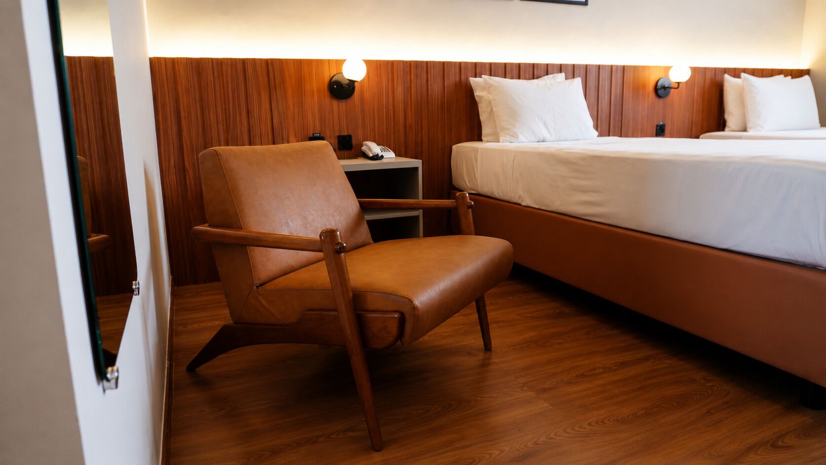 A hotel room with a wooden floor, a bed with white bedding, a brown leather chair, a bedside table with a lamp, and a phone.