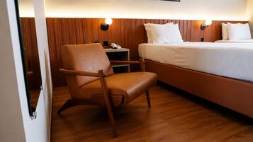 A hotel room with a wooden floor, a bed with white bedding, a brown leather chair, a bedside table with a lamp, and a phone.