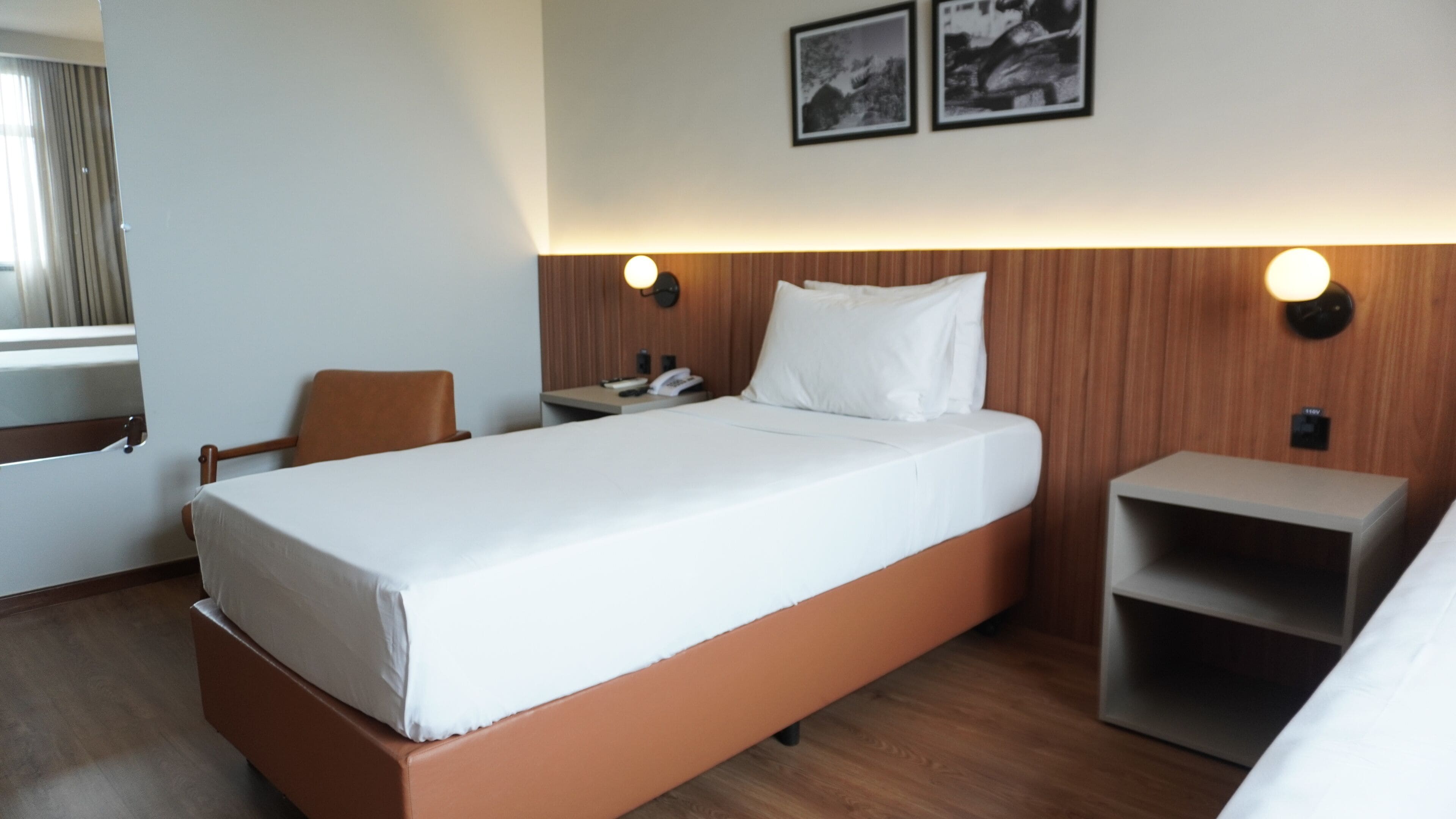 A hotel room with a single bed, a chair, a bedside table, a nightstand, and two wall-mounted lights.