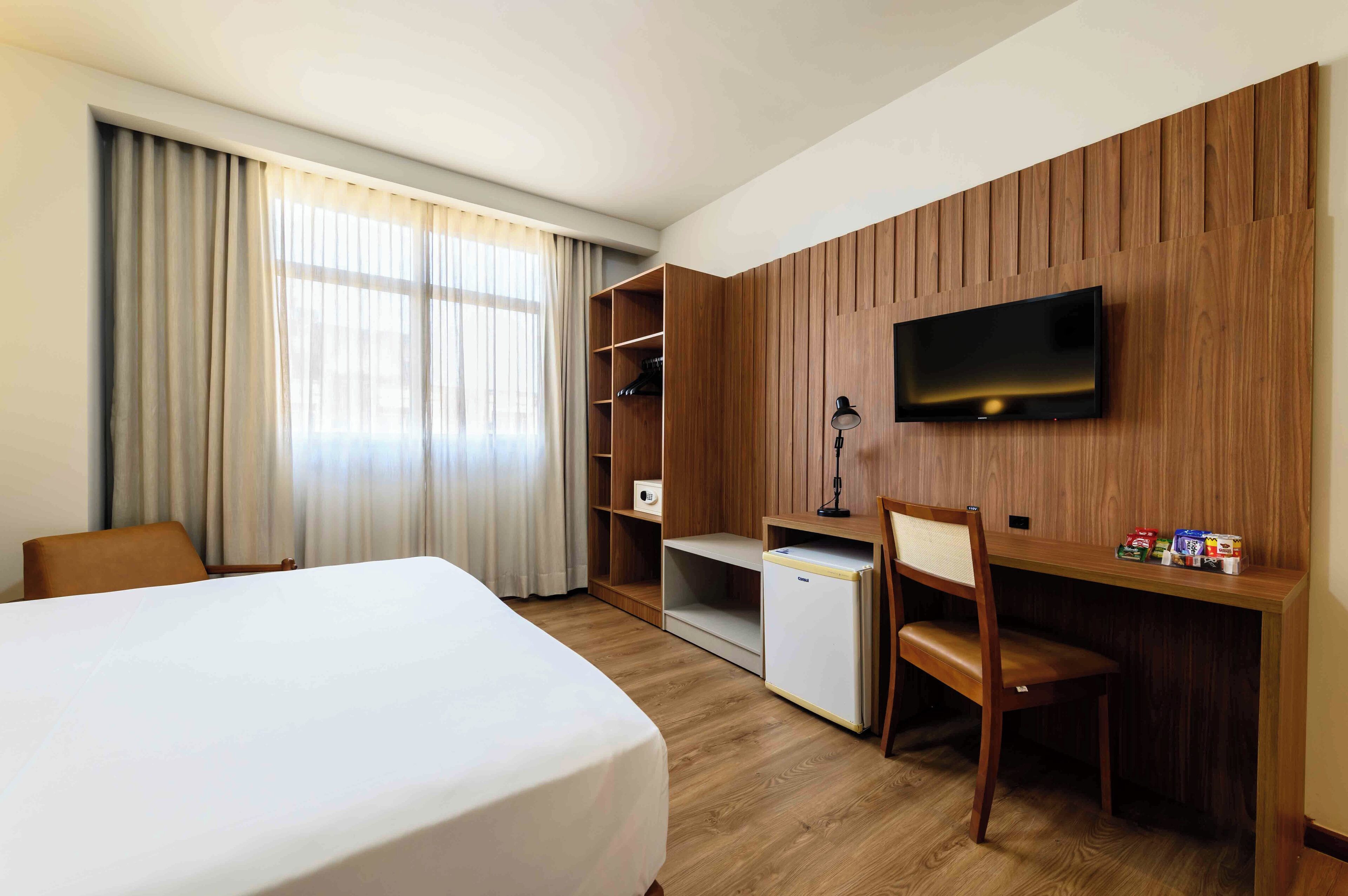 Habitación de hotel con cama, escritorio con silla, televisor montado en la pared y armario con estantes.