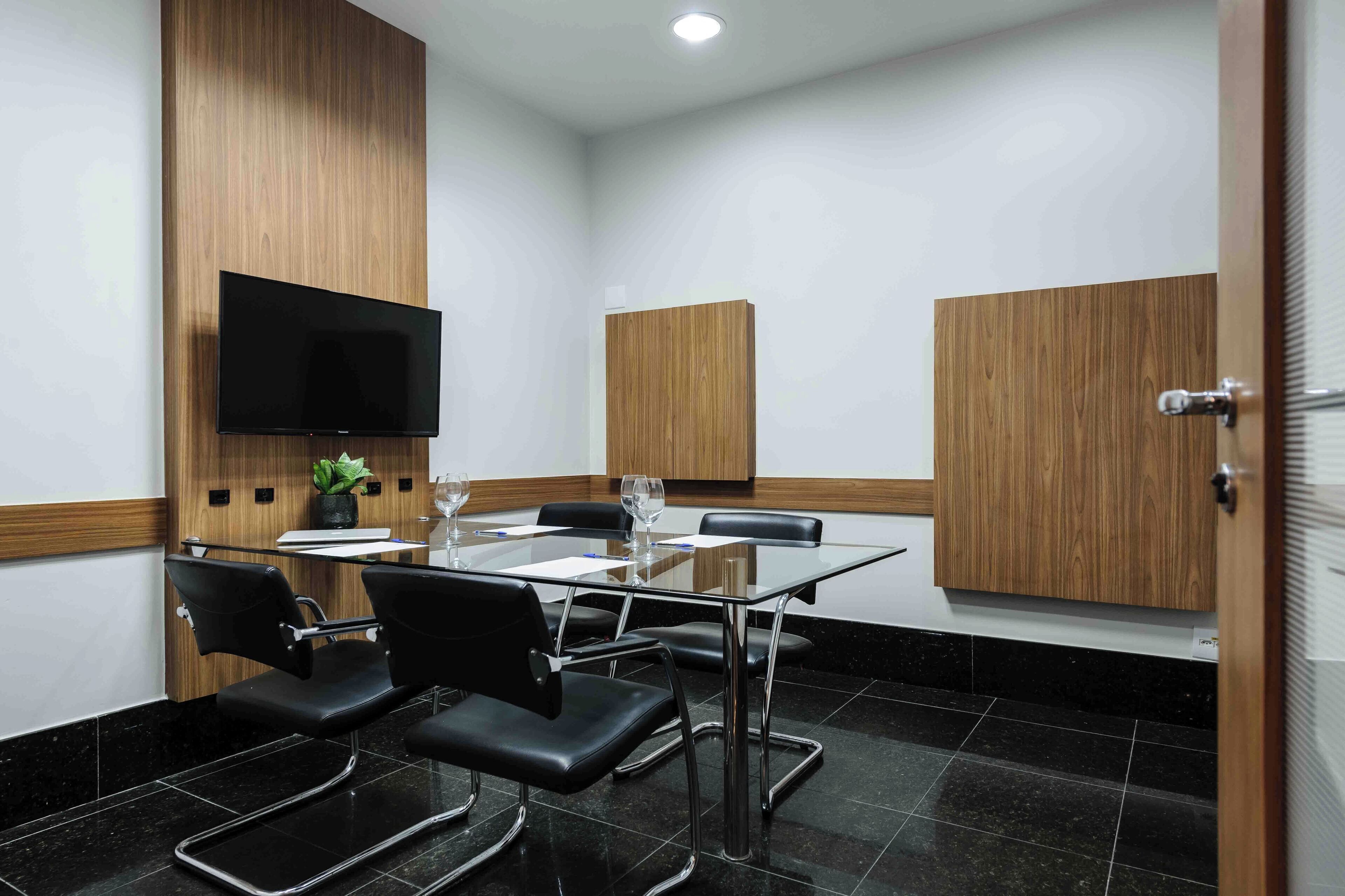 Un salón de conferencias con una mesa de vidrio, sillas negras, un televisor montado en la pared y paneles de madera.