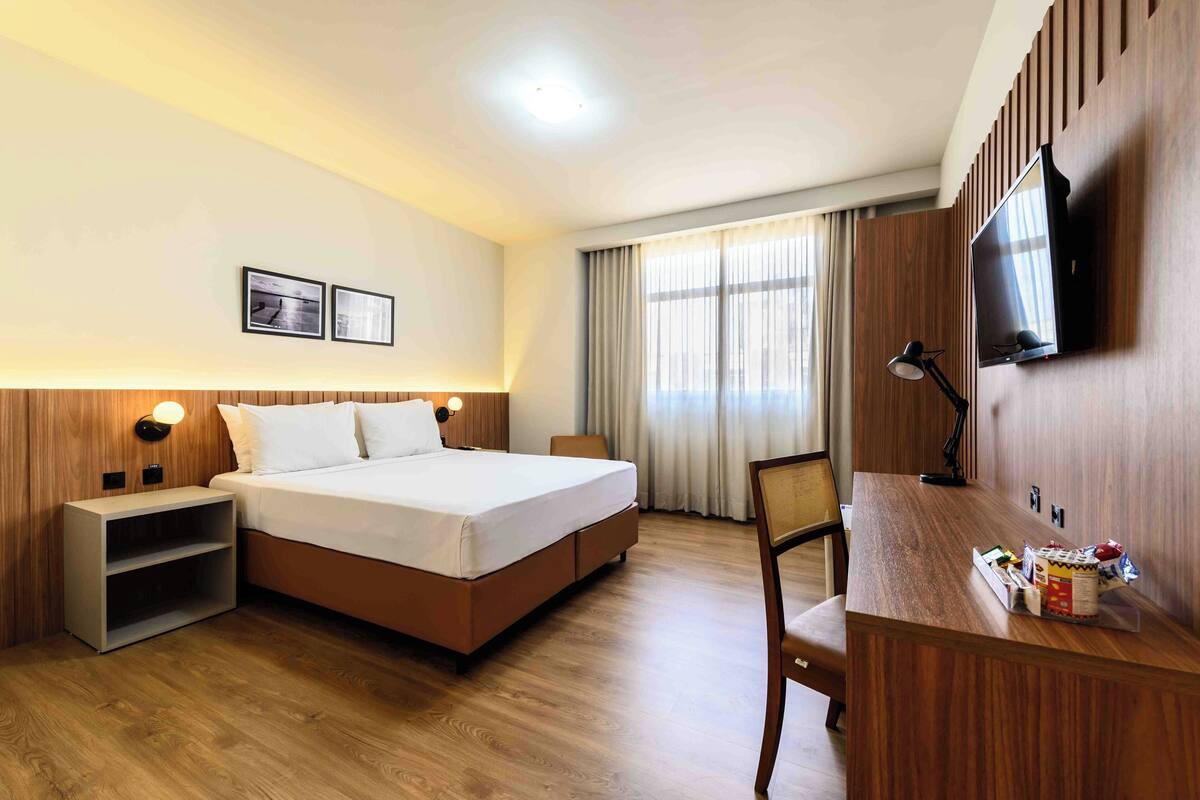 Une chambre d’hôtel comprenant un lit, des tables de chevet, un bureau, une chaise, une télévision et une fenêtre avec des rideaux.