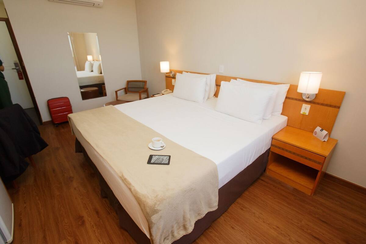 Une chambre d’hôtel avec un lit, des tables de chevet, une chaise, un bureau, un miroir et une valise rouge.
