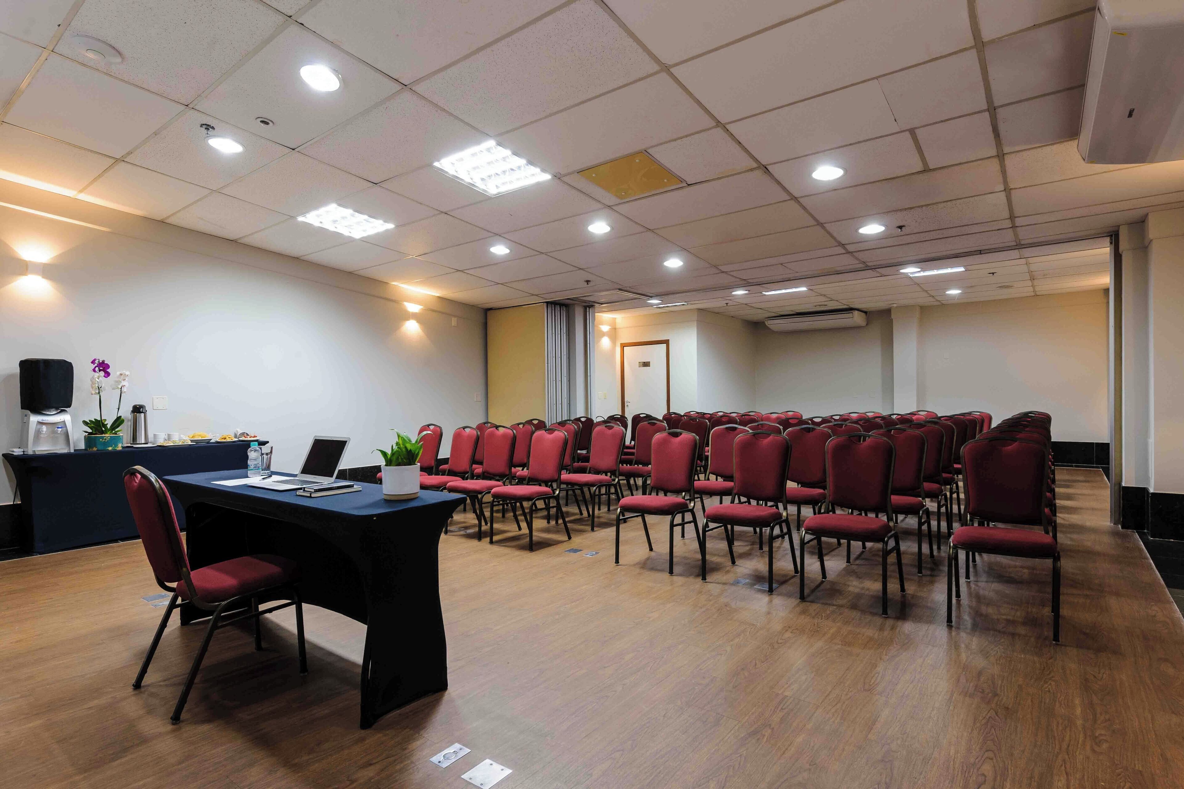 Un salón de conferencias con filas de sillas rojas, una mesa negra con un ordenador portátil y un mostrador de recepción con plantas.