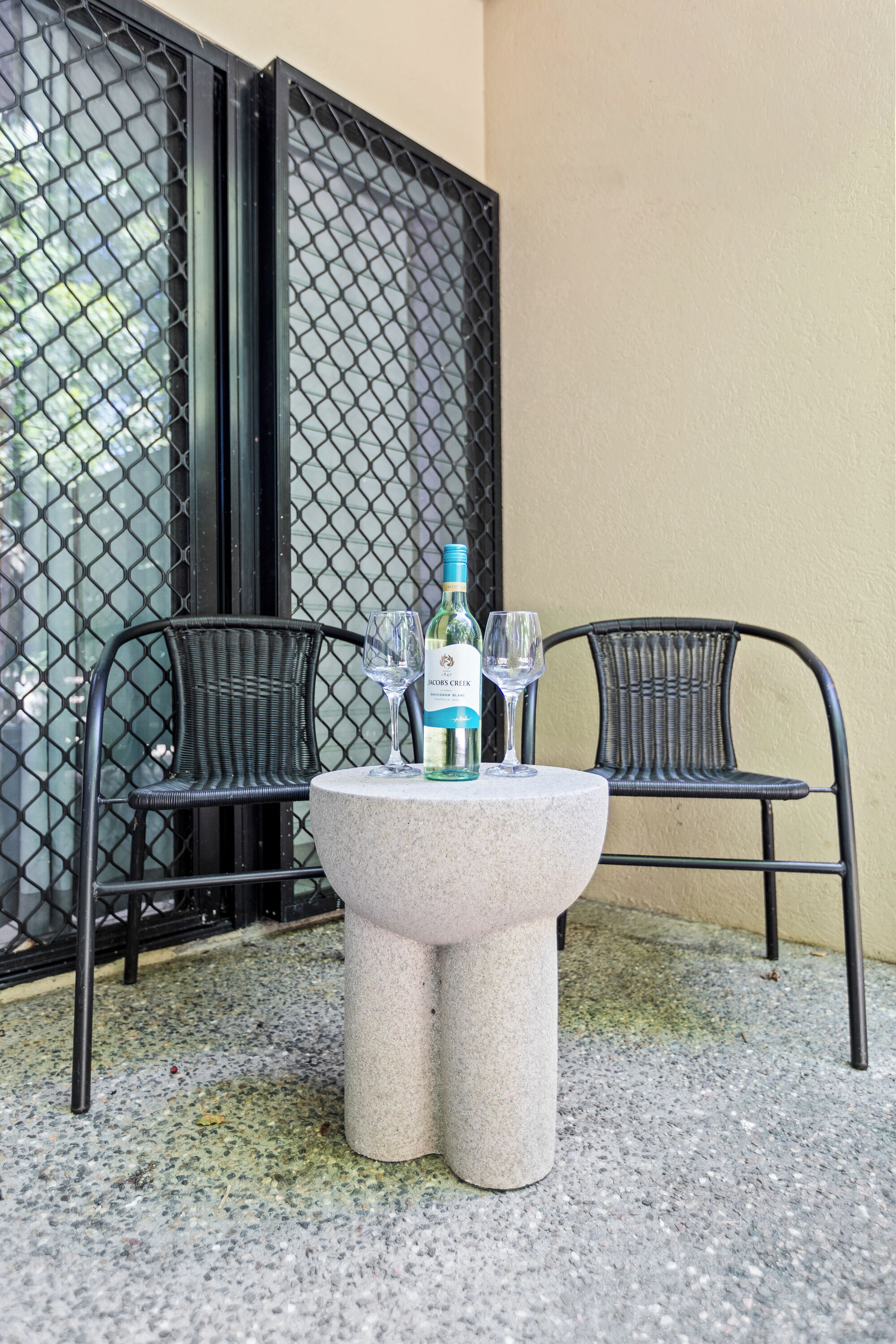 A table with a bottle and two glasses, two wicker chairs, and a metal fence.