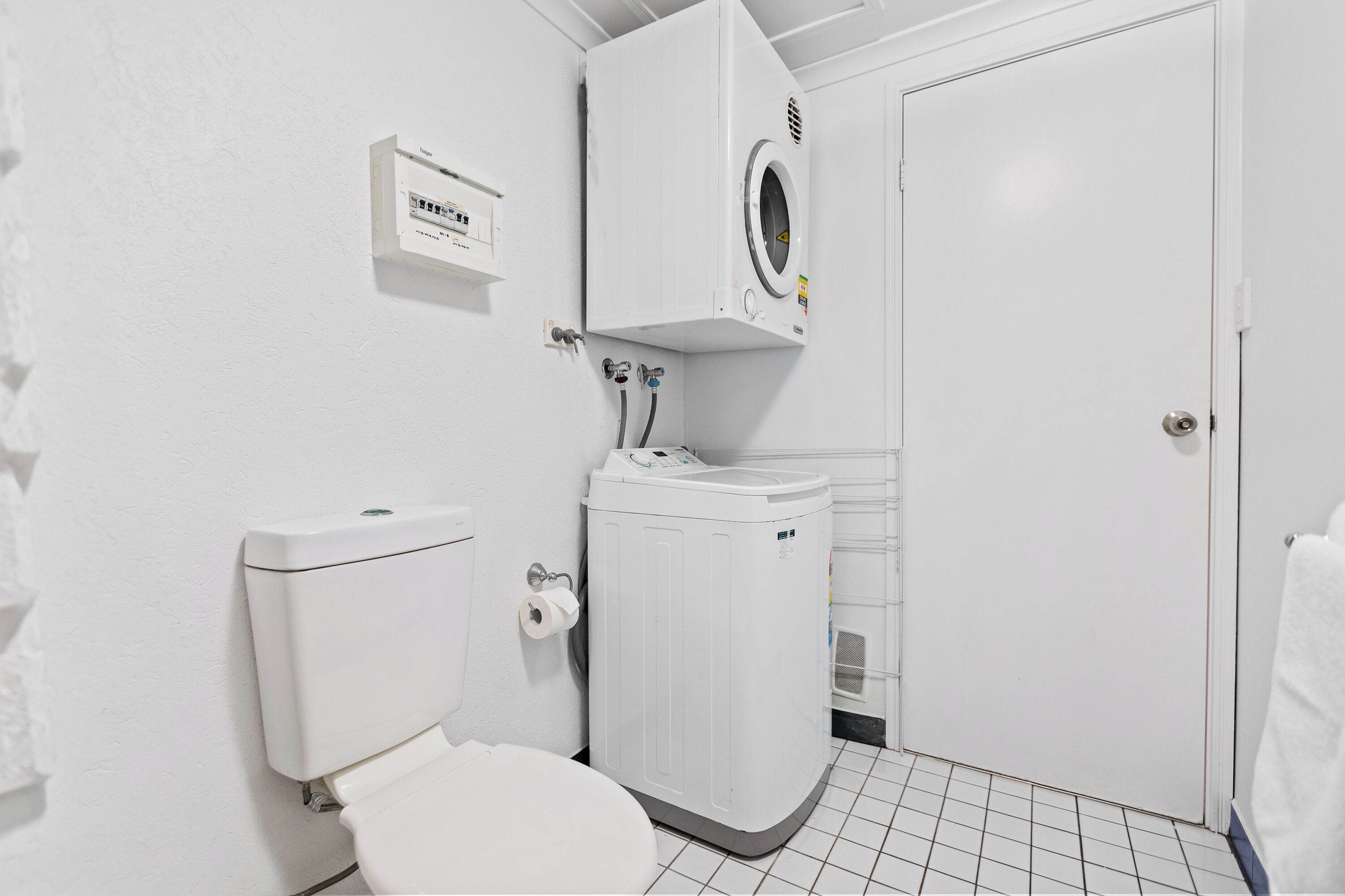 A compact, white-walled bathroom with a toilet, a washing machine, and a small electrical outlet on the wall.