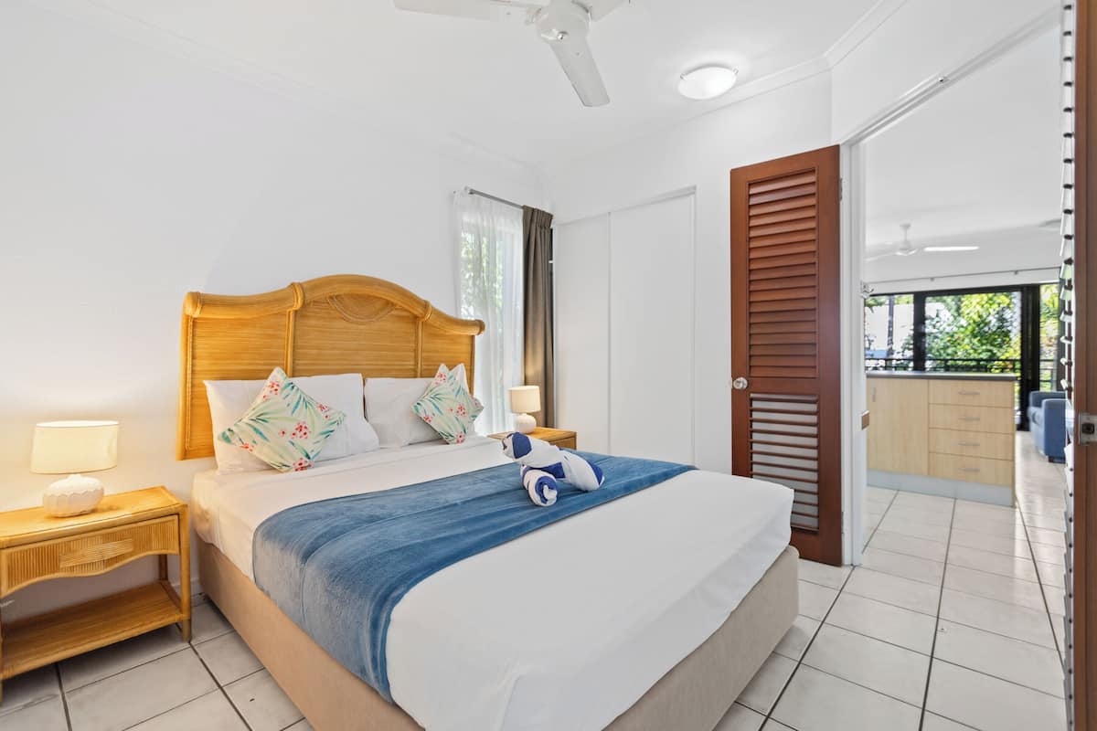 A bedroom with a bed, wooden headboard, bedside tables with lamps, a blue blanket, and a view of an outdoor area through a door.