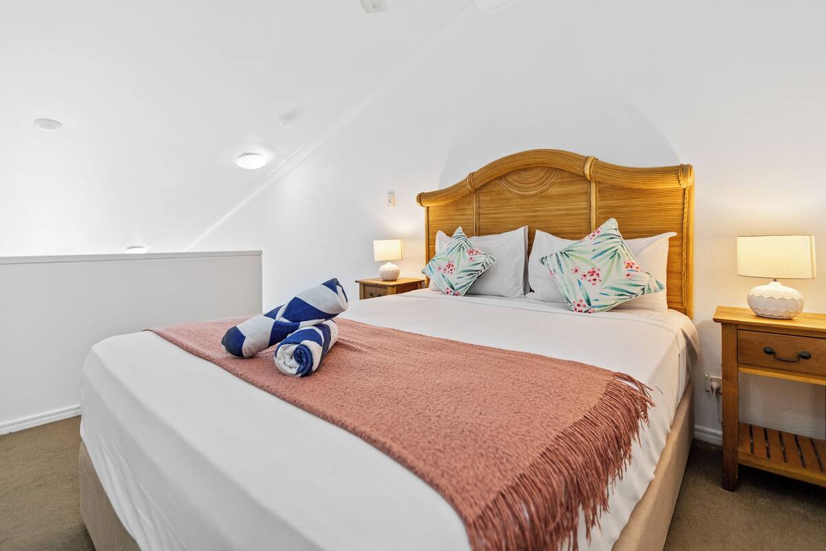 A neatly made bed with a wooden headboard, a pink throw blanket, and a blue and white towel. There are two bedside tables with lamps and a wall-mounted light.