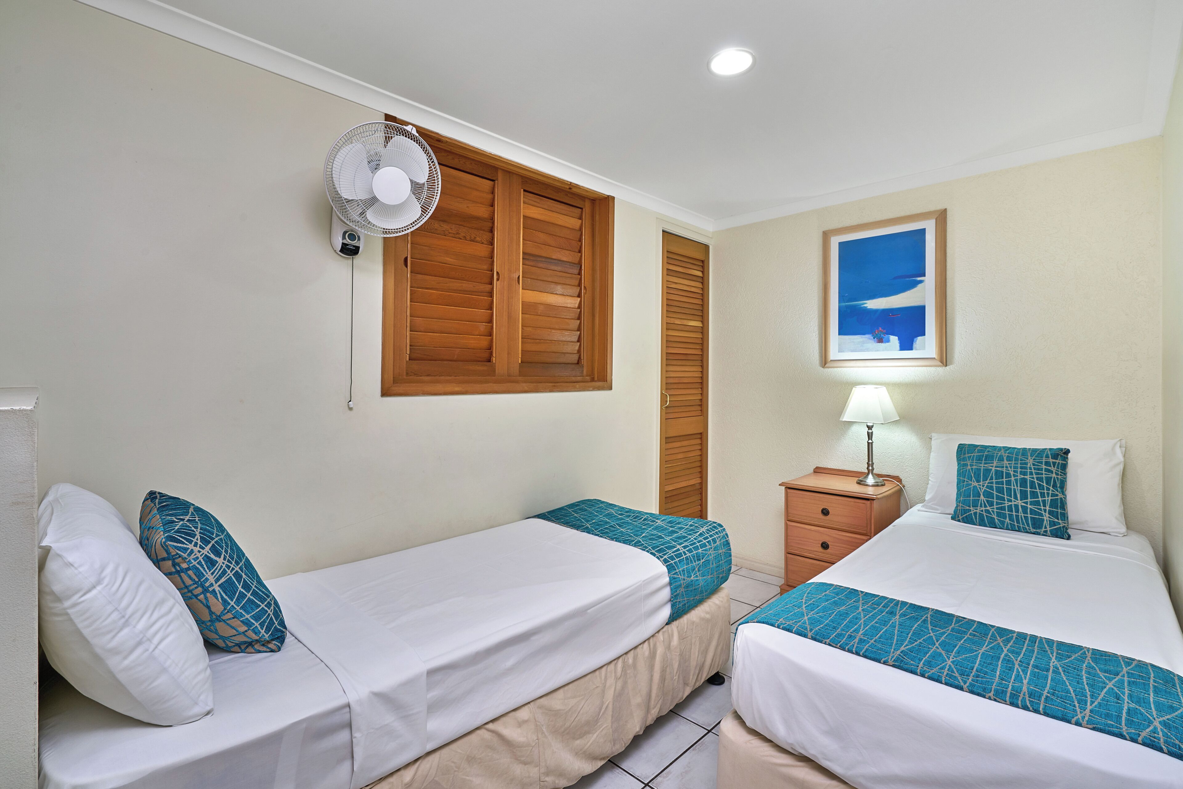 A hotel room with two single beds, a bedside table with a lamp, a wall-mounted fan, and a window with wooden shutters.
