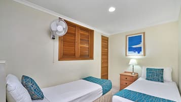 A hotel room with two single beds, a bedside table with a lamp, a wall-mounted fan, and a window with wooden shutters.
