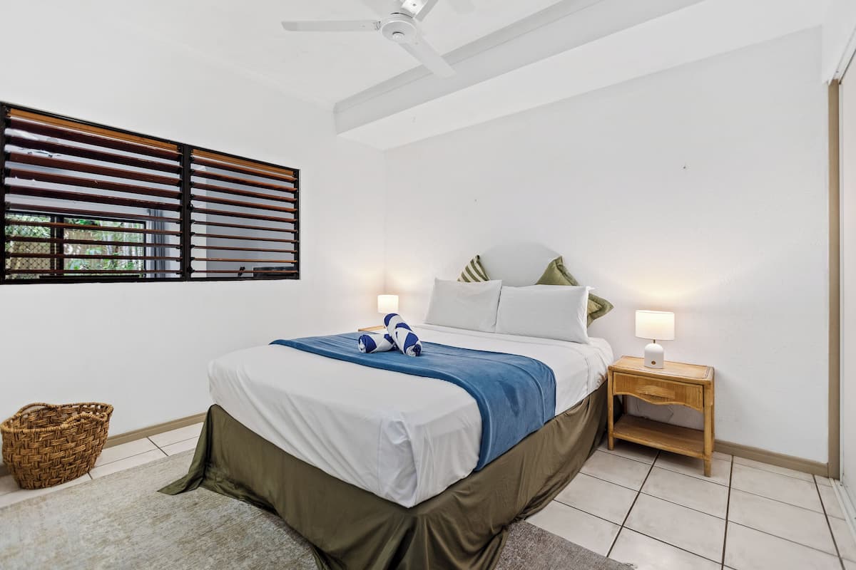 A bedroom with a bed, bedside table, lamp, and a window with blinds.