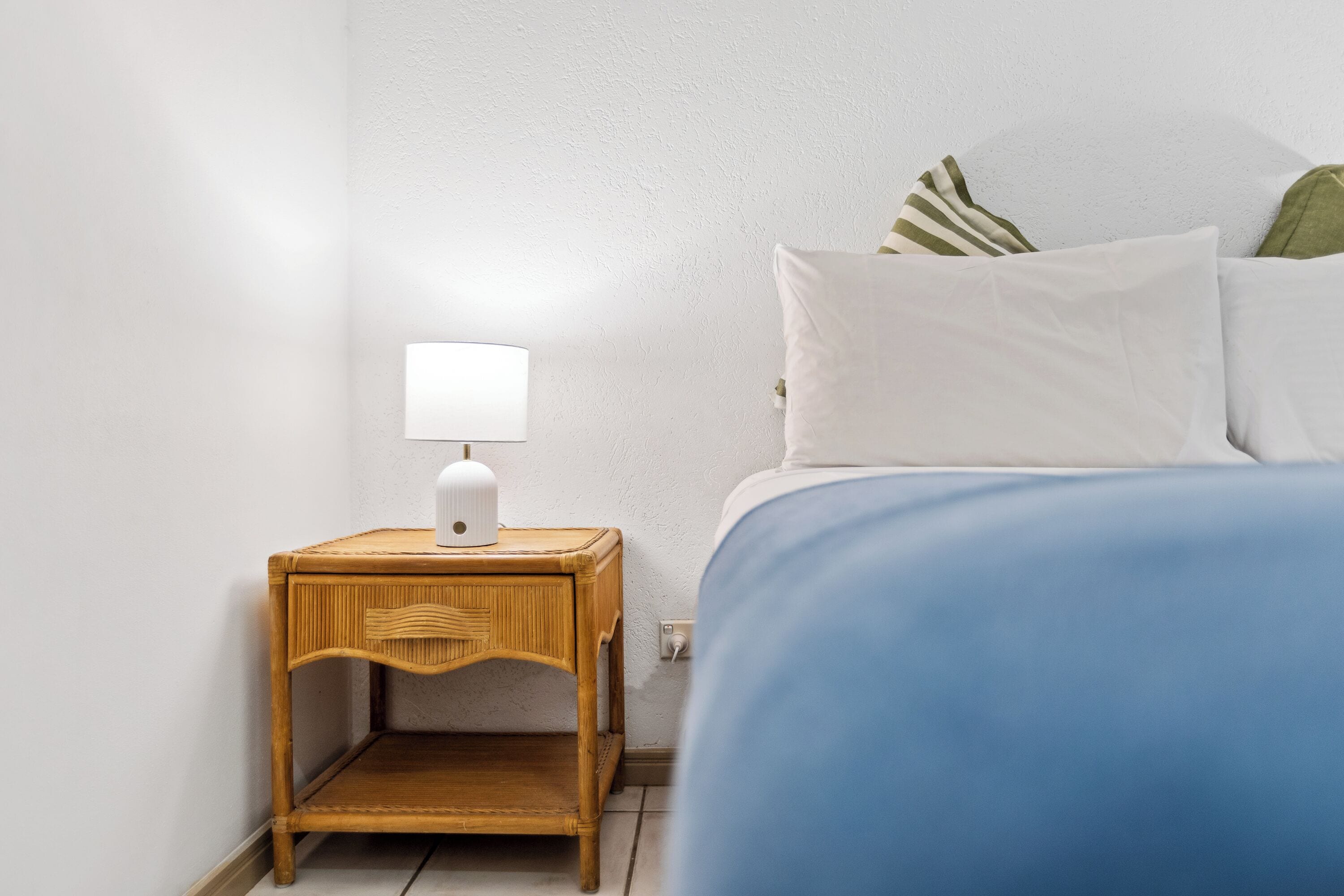 A neatly made bed with a blue cover, a small wooden nightstand with a white lamp, and a white wall.
