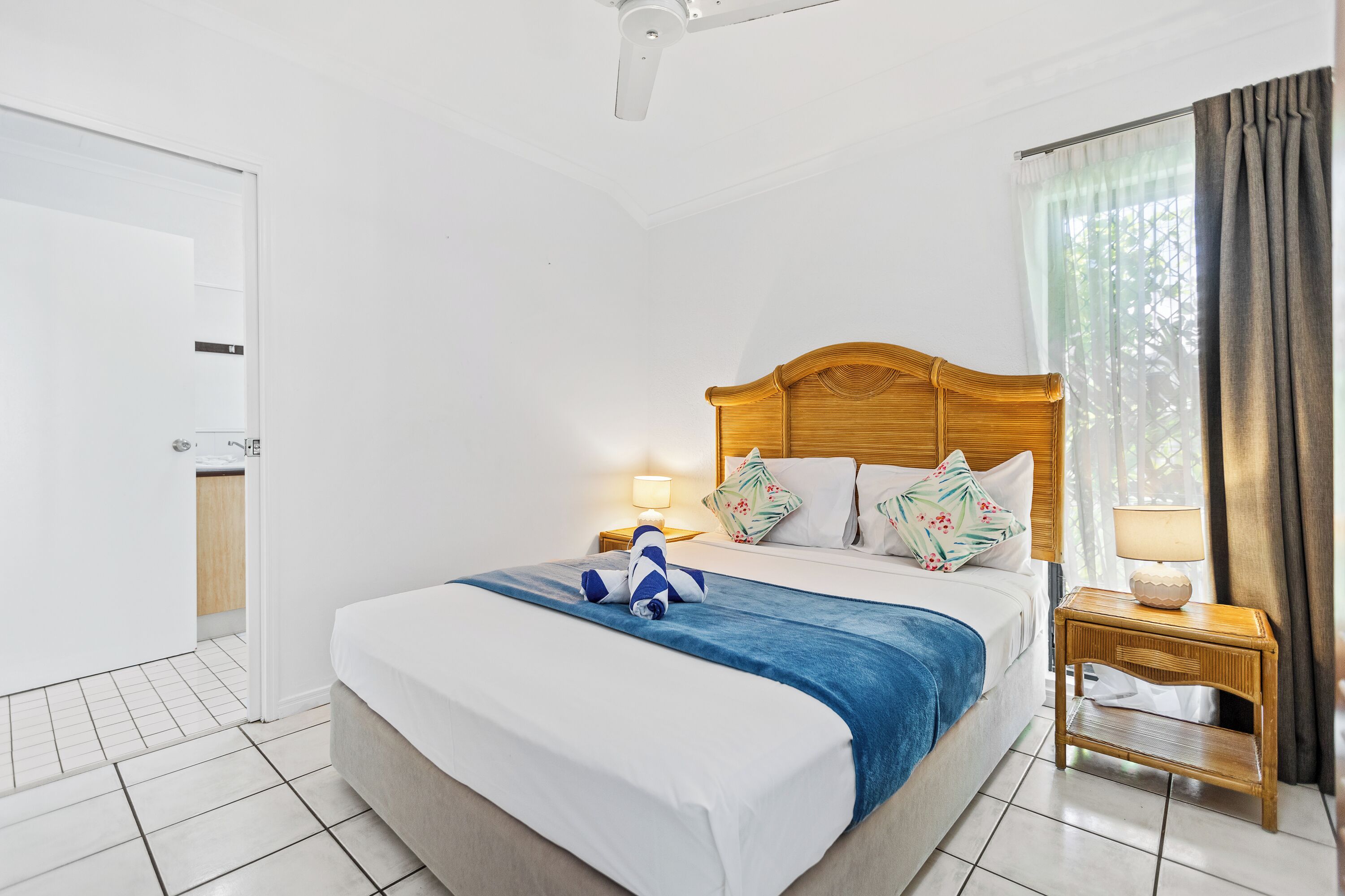Una habitación de hotel con cama, mesita de noche, lámpara, ventilador de techo y ventana con cortinas.