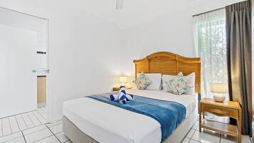 Une chambre d’hôtel avec un lit, une table de chevet, une lampe, un ventilateur de plafond et une fenêtre avec des rideaux.