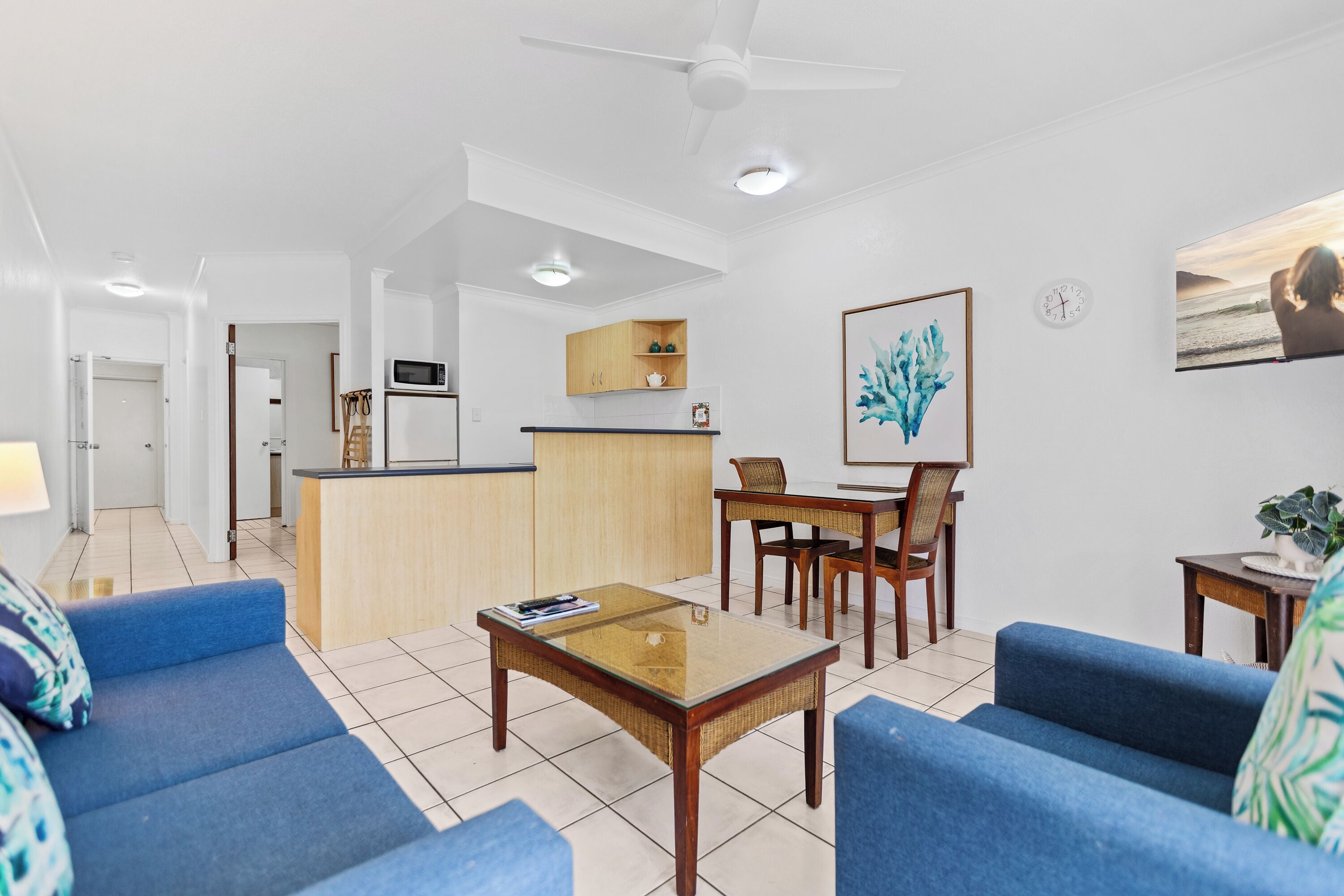 A modern living room with a glass coffee table, blue sofas, a dining area with wooden chairs, a kitchen with a microwave, and a ceiling fan.