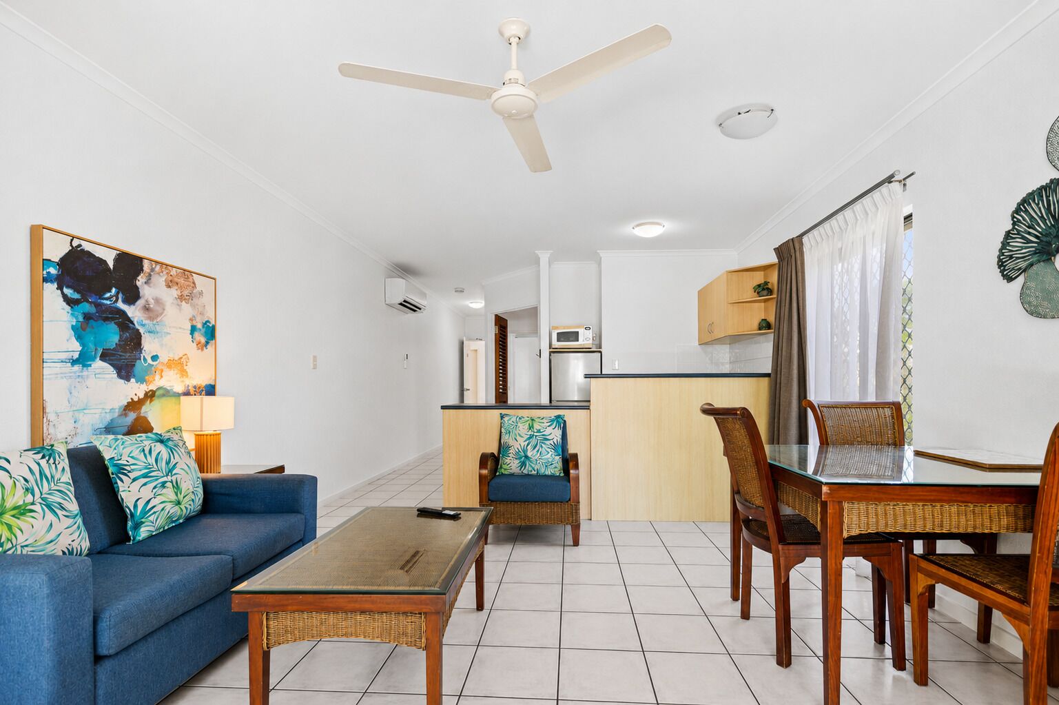 A living room with a sofa, coffee table, dining table, chairs, a ceiling fan, and a kitchen area in the background.