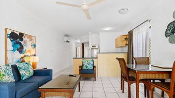 A living room with a sofa, coffee table, dining table, chairs, a ceiling fan, and a kitchen area in the background.