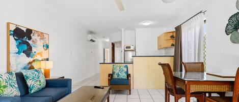 Una sala de estar con sofá, mesa de centro, mesa de comedor, sillas, ventilador de techo y una zona de cocina al fondo.