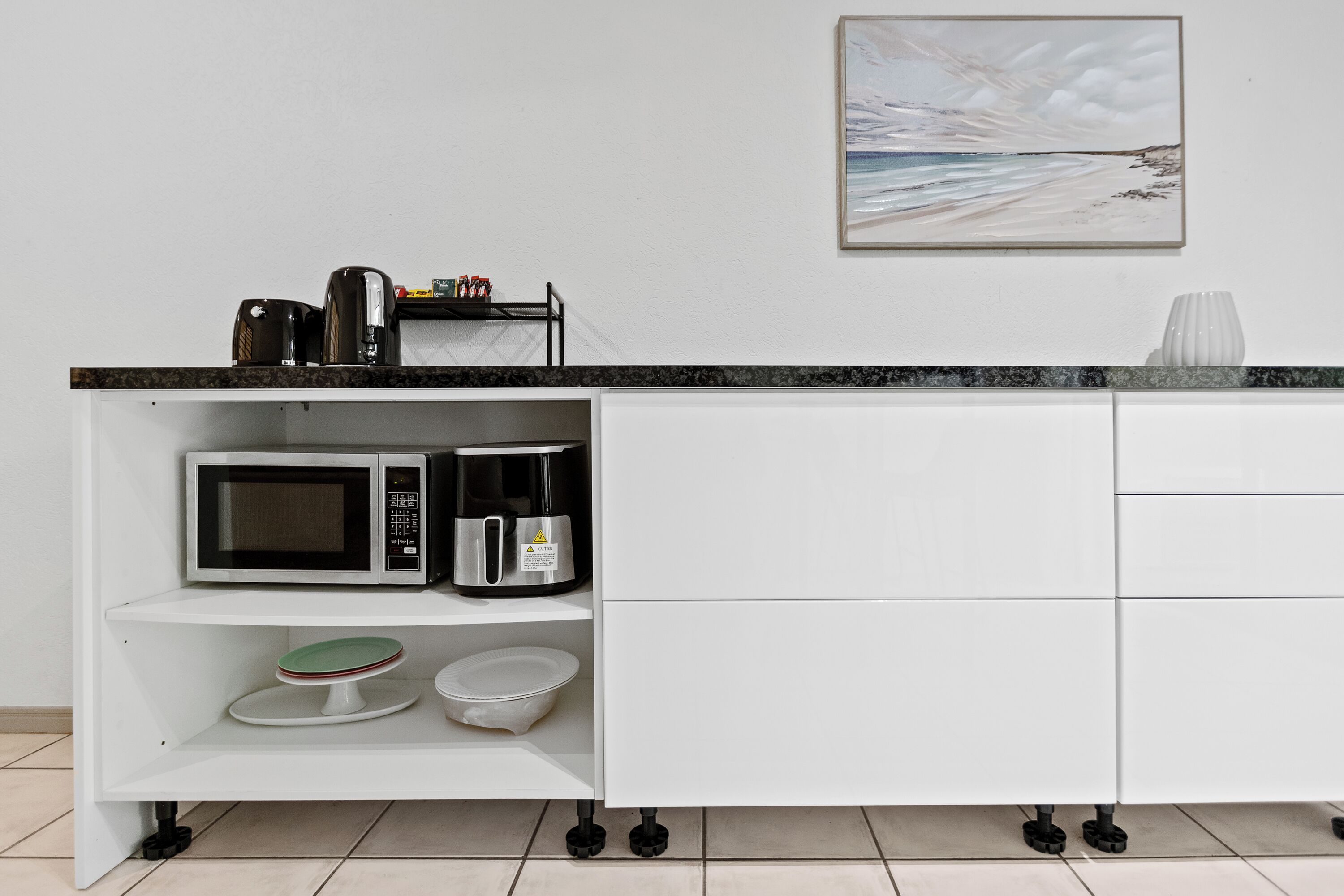 Una cocina con microondas, cafetera y una estantería con vajilla.