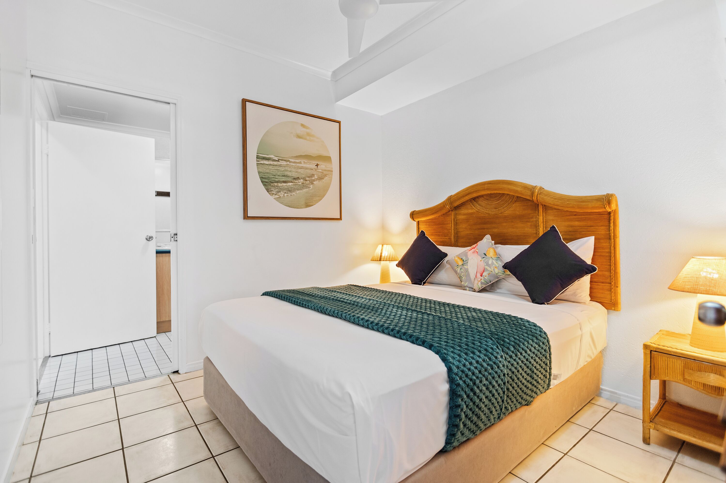 A neatly made bed with a wooden headboard, a green blanket, and multiple pillows. There is a framed picture on the wall, a bedside lamp, and a small wooden nightstand.