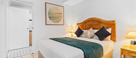 A neatly made bed with a wooden headboard, a green blanket, and multiple pillows. There is a framed picture on the wall, a bedside lamp, and a small wooden nightstand.