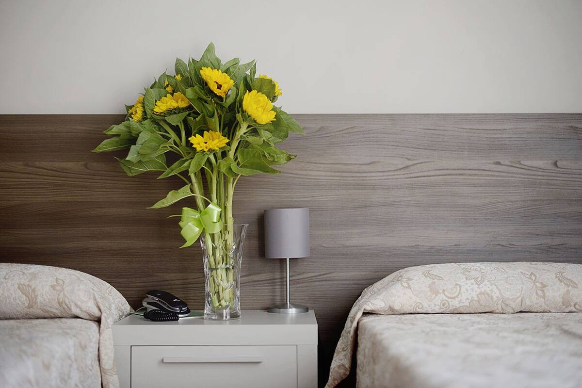 A hotel room with a wooden headboard, a bedside table with a lamp, a vase with yellow flowers, and a telephone.